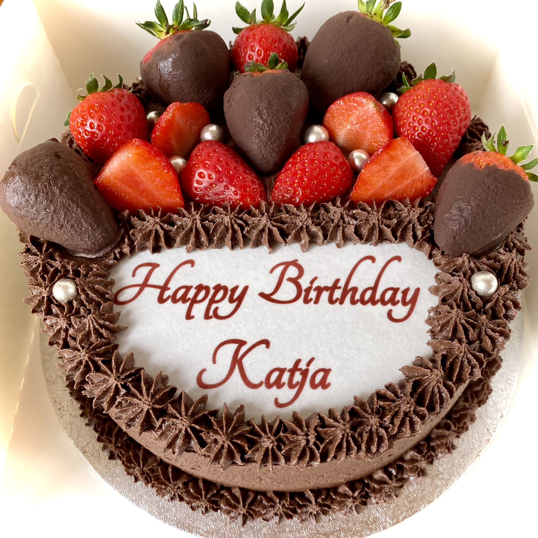 A decadent chocolate cake adorned with fresh strawberries and chocolate-dipped strawberries. The cake features a heartfelt message in red icing, 'Happy Birthday Katja,' displayed on a white icing plaque, surrounded by intricate chocolate piping and shimmering silver edible pearls. A perfect dessert for any celebration!